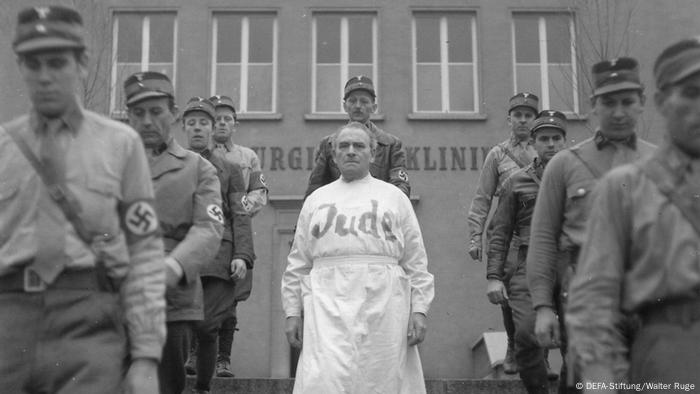 Filmstill aus Professor Mamlock, mit PM, der in Arztkittel mit Aufschrift Jude zwischen Nazis abgeführt wird (DEFA-Stiftung/Walter Ruge)