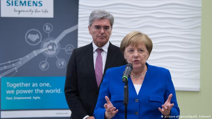 Merkel speaks in front of Siemens chairman Joe Kaeser as they stand in front of a Siemens sign