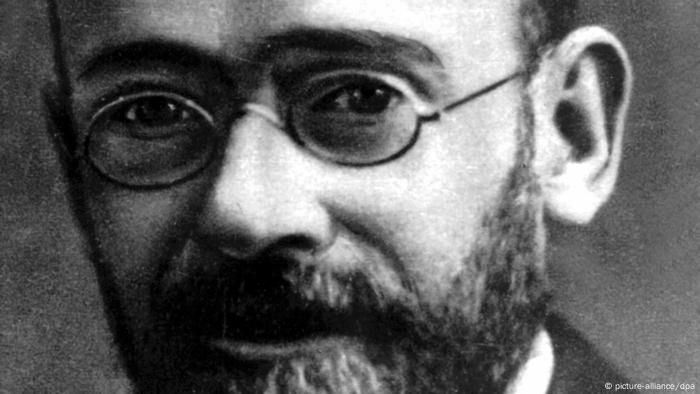 Black and white photo in tight close-up of a man with sad, dark eyes, oval glasse,s and a full, dark beard.