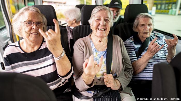 Drei rüstige Rentnerinnen sitzen in einem Bus und zeigen das Wacken- bzw. Metalsymbol, die Pommesgabel oder Teufelshörner, mit Zeigefinger und kleinem Finger