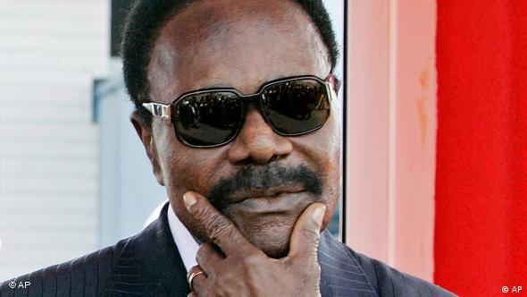 Former President of Gabon Omar Bongo faces the camera. He is wearing black sunglasses and holding his thumb and forefinger to his chin.