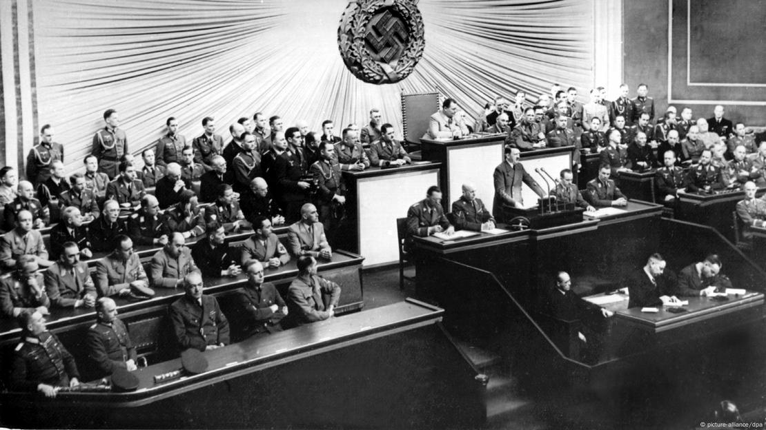 Hitler en la tribuna de oradores del Reichstag, con representantes del gobierno nazi sentados a su lado y detrás de él bajo una esvástica.