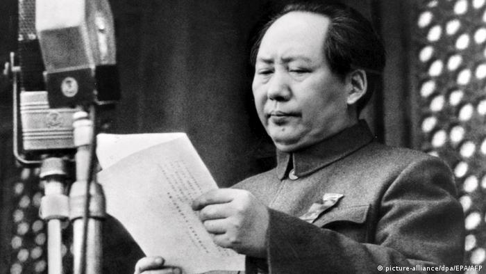 Ein schwarz-weiß Foto von Mao Zedong, der ein Blatt Papier in der Hand hält und vor zwei Mikrofonen steht