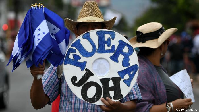 Foto de una persona con un rótulo que dice Fuera JOH y banderas de Honduras en la mano.