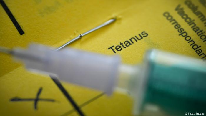 Desfocada, em primeiro plano, está uma seringa de vacinação. Atrás, um papel amarelo com a palavra Tetanus. 