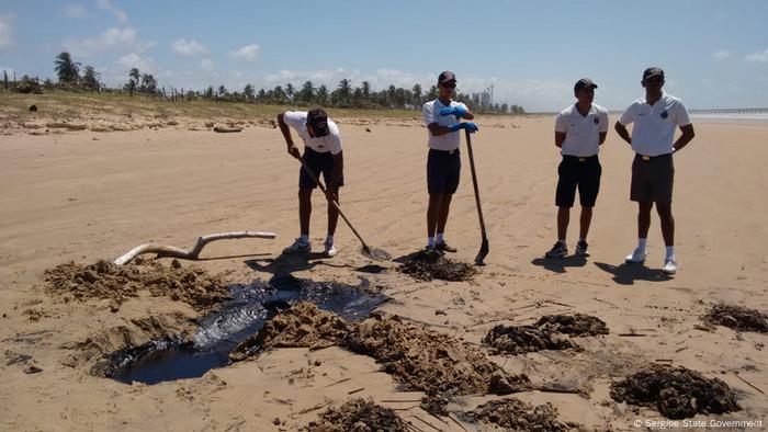 Men with shovels stand in front of a beach oil spill in Brazil