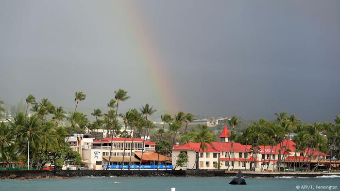 Ein Regenbogen lässt die feuchtwarme Luft über dem Ort Kailua Kona erahnen, dessen rote Dächer durch viele grüne Palmen hindurchleuchten. Im Vordergrund schwappt der Pazifik an die schwarze Kaimauer aus Lavasteinen. Ein Regenbogen lässt die feuchtwarme Luft über dem Ort Kailua Kona erahnen, dessen rote Dächer durch viele grüne Palmen hindurchleuchten. Im Vordergrund schwappt der Pazifik an die schwarze Kaimauer aus Lavasteinen.