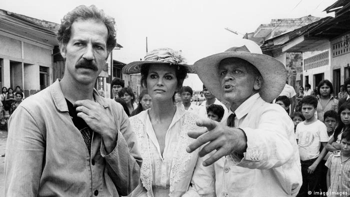 Director Werner Herzog (left) with actors Claudia Cardinale and Klaus Kinski in period costumes amid curious children from a South American village.