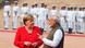 Merkel and Modi talk to each other. Merkel is wearing red and Modi is wearing a brown waistcoat and white shirt. Presidential guards stand in the background Merkel and Modi talk to each other. Merkel is wearing red and Modi is wearing a brown waistcoat and white shirt. Presidential guards stand in the background