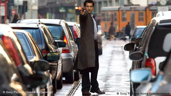 Mann mit gezogener Pistole auf einer Straße zwischen parkenden Autos - Szene aus The International