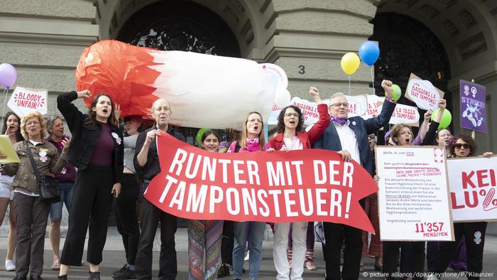 People protesting holding a signs that says lower the tampon tax with a large fake tampon in the background