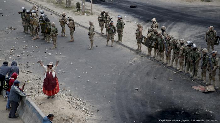 Mujer indígena alza los brazos en el medio de una calle flanqueada por militares, durante una protesta en El Alto