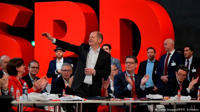 Berlin | SPD-Bundesparteitag im Dezember 2019. Olaf Scholz steht an seinem Platz, hat den Arm winkend gehoben und lächelt. Die um ihn herum sitzenden Delegierten applaudieren