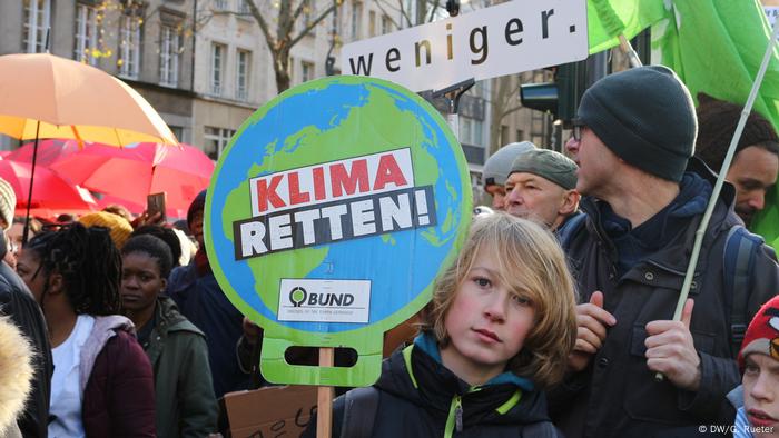 Demonstration von Fridays for Future. Ein Junge hält ein Plakat. Auf dem steht: Klima Retten. Dahinter hält ein Mann ein Plakat: Darauf steht: Weniger 