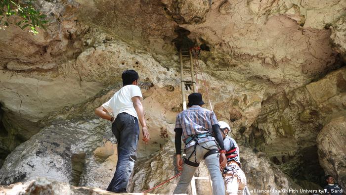 Drei Männer stehen vor dem Eingang zu einer Höhle, zu dem eine Leiter hinaufführt. aEiner von ihnen trägt einen Helm