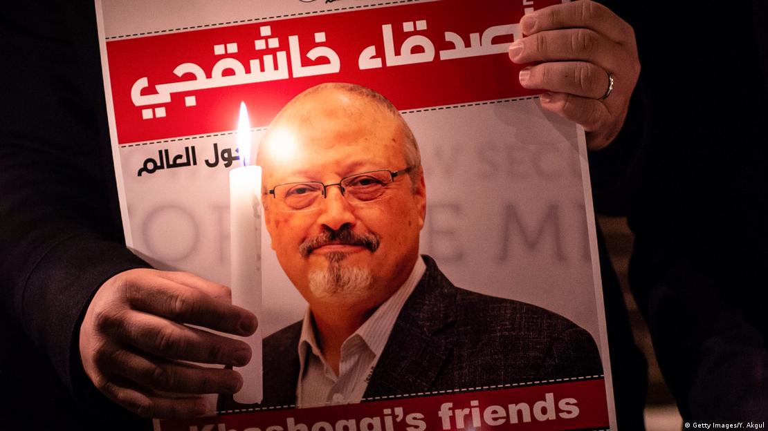 Yüzü görünmeyen bir kişi bir elinde gazeteci Cemal Kaşıkçı posterini, diğer elinde ise yanan bir mum tutuyor. Yüzü görünmeyen bir kişi bir elinde gazeteci Cemal Kaşıkçı posterini, diğer elinde ise yanan bir mum tutuyor.