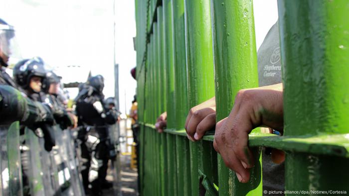 Migrantes detrás del muro fronterizo entre México y Guatemala, frente a policías armados.