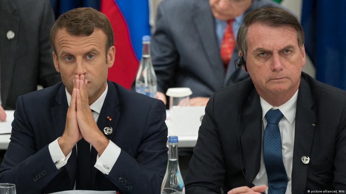 Macron com as mãos sobre a boca sentado ao lado de Bolsonaro, sério