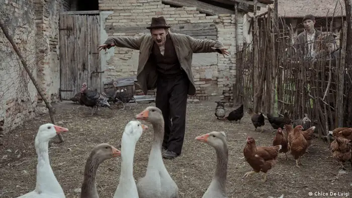 Ein Mann mit Schnurrbart und Hut steht in einem Stall mit Gänsen und Hühnern und macht eine Geste wie eine Vogelscheuche (Chico De Luigi)