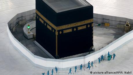 The imposing black square building of the Kaaba sits in the center of the Grand Mosque in Mecca while a line of seemingly tiny men in green sanitation uniforms clean the usually busy floor of white tiles