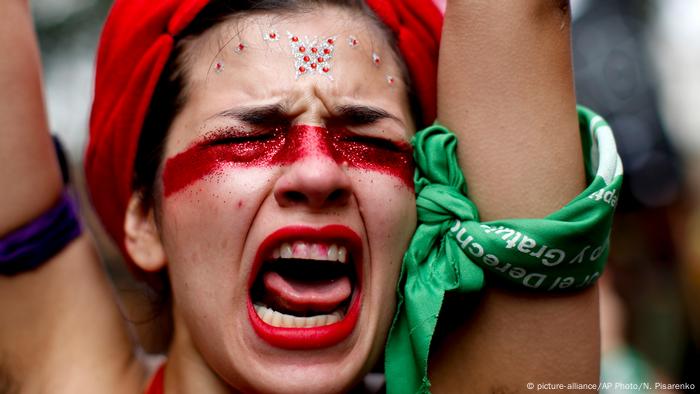 Una mujer con la boca abierta, los ojos cerrados, y un pañuelo verde en un brazo levantado.