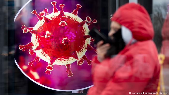 A person wearing a mask stands in front of an image of the novel coronavirus in Berlin