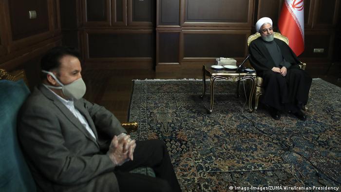 President Rouhani sits in a chair while a minister wears a mask