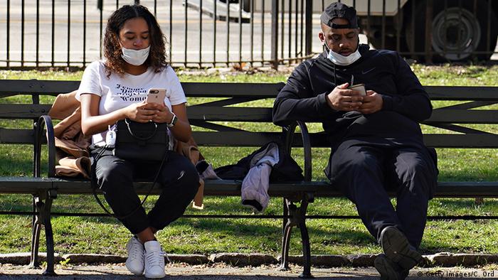 Two people sitting on a bench wearing protective masks using their phones