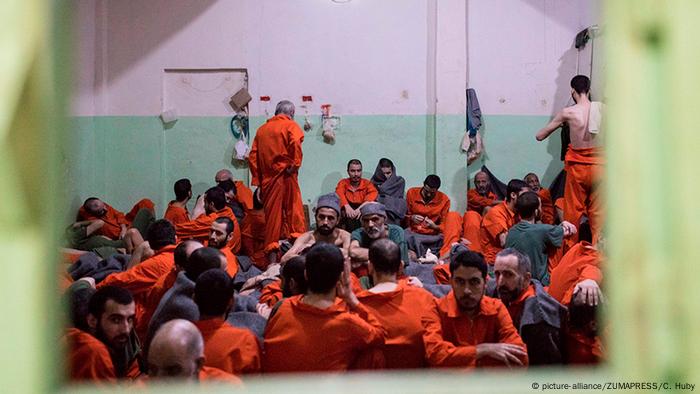 Men wearing orange jumpsuits crowded together in a room, sitting on the floor