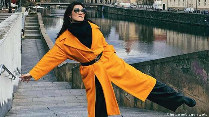 Olga Peretyatko in a yellow coat, black sweater and large sunglasses, alone on concrete steps next to a river in a city