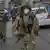 A mask-wearing soldier carrying a gun walks down a street