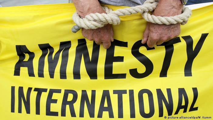 An Amnesty International activist with his hands fake tied by a rope holds a banner with the organization's name