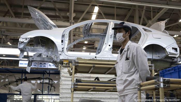 Homem de boné, máscara e roupa branca em uma linha de montagem. Atrás, um carro branco sendo montado. 