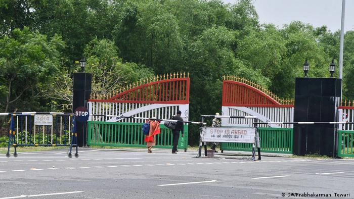 Two people walk through guarded gate painted with colors of Indian flag. In front of gate is a long barrier and signs saying stop
