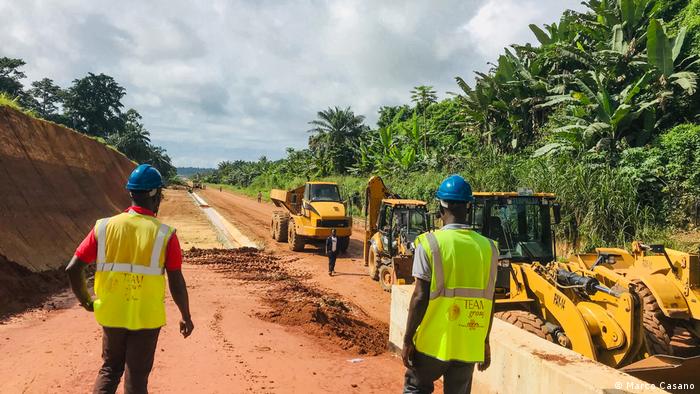 Workers in Kojokrom, Takoradi, stand on a cleared strip of red dirt look at earth-moving equipment 