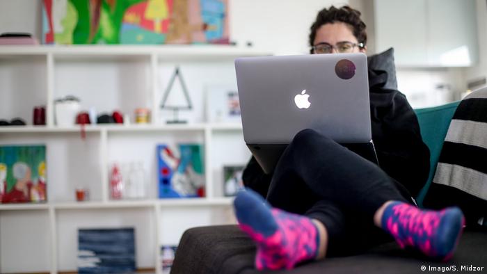 A woman in comfortable clothes and bright pink socks works on an Apple laptop on a sofa.