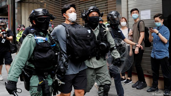 Zwei Polizisten mit Helm und Schutzausrüstung führen einen jungen Mann ab, der einen Nase-Mund-Schutz und einen Rucksack vor der Brust trägt; Proteste in Hongkong