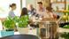 A cooking pot sits on the stovetop next to fresh herbs while in the background a family sits around a table A cooking pot sits on the stovetop next to fresh herbs while in the background a family sits around a table