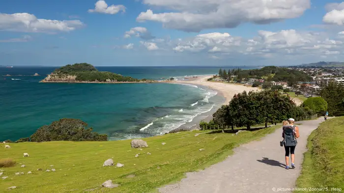 A hiker carries a baby on her back along a nature trail with the ocean and a town in the background