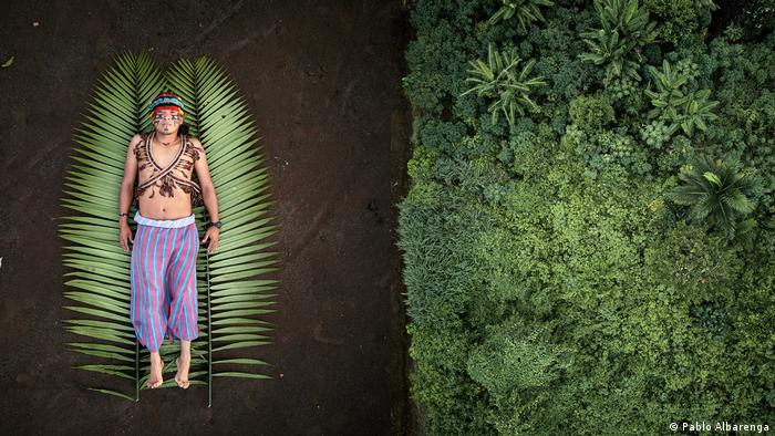 A man wearing face paint and traditional dress lies on a two palm leaves. His image juxtaposed with an aerial view of a forest