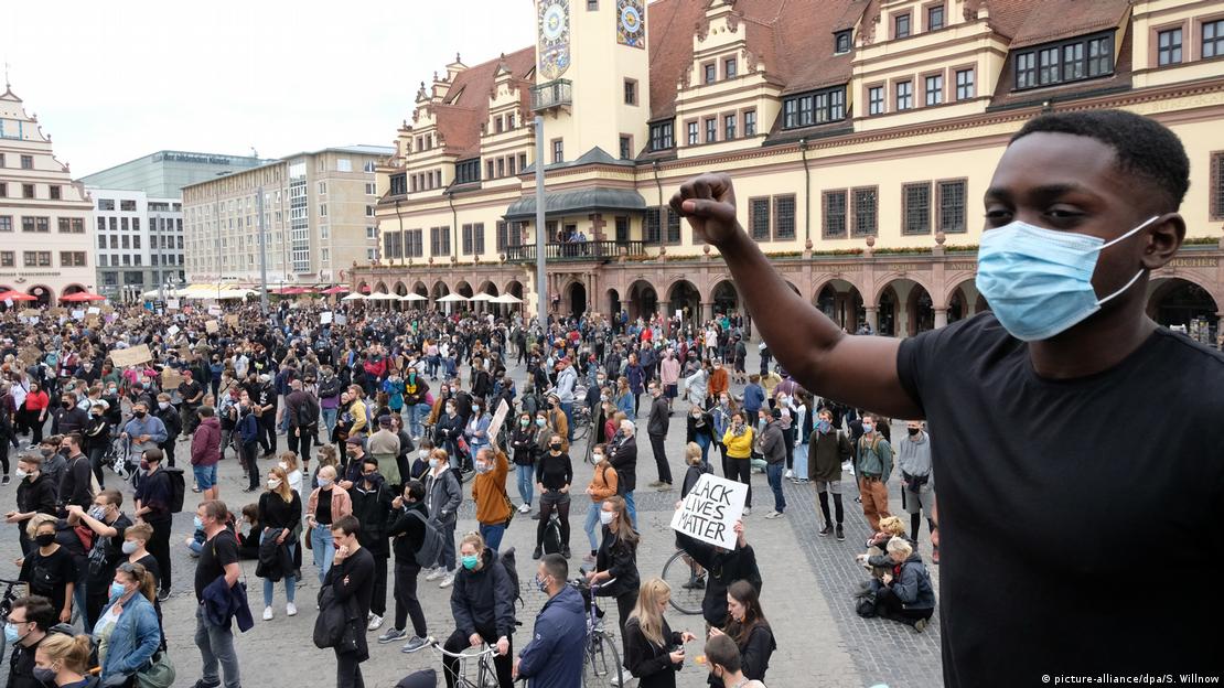 Foto externa mostra em primeiro plano rapaz negro com o punho erguido. Ao fundo, pessoas em manifestação em cidade na Alemanha.