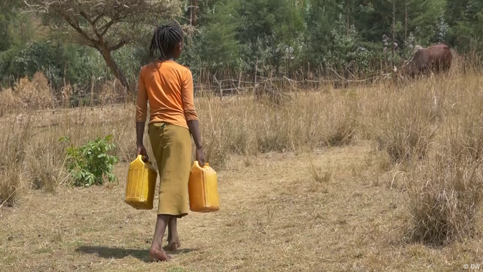 A woman carrying two water canisters walks through a dry landscape