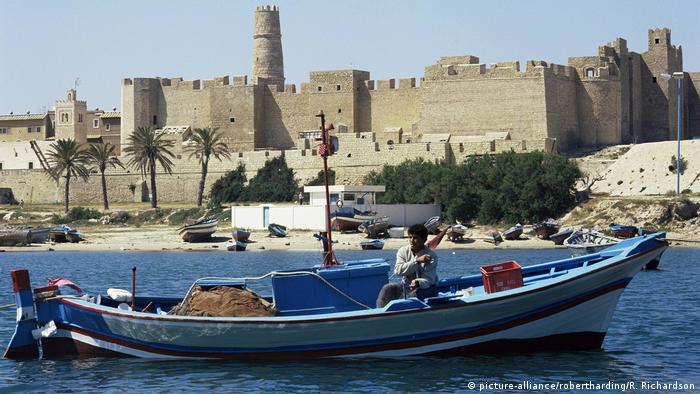 A man sits in a small boat on a stretch of water with a towering fortress in the background