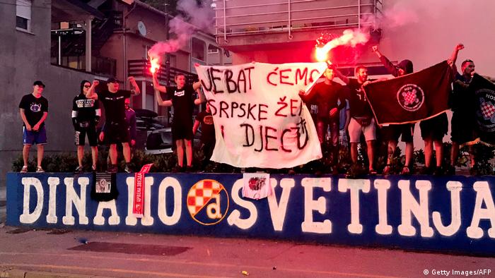 Rechtsextreme Fans des Zagreber Fußballclubs stehen auf einer Bühne und halten ein Transparent, auf dem eine brutale, vulgäre, sexistische und serbenfeindliche Parole steht, zwei Männer halten Fackeln 