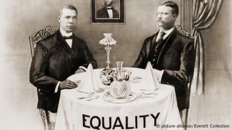 Booker T. Washington und Präsident Roosevelt sitzen an einem Tisch, auf dem Tischtuch steht Equality