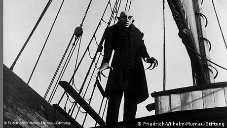 Count Orlok stands on a ship's deck. He has bat-like ears, thick-eyebrows and long canines.