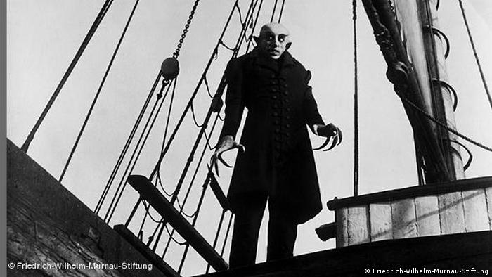 Count Orlok stands on a ship's deck. He has bat-like ears, thick-eyebrows and long canines.