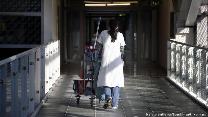 Woman cleaner in white jacket pushing cleaning cart along a hall
