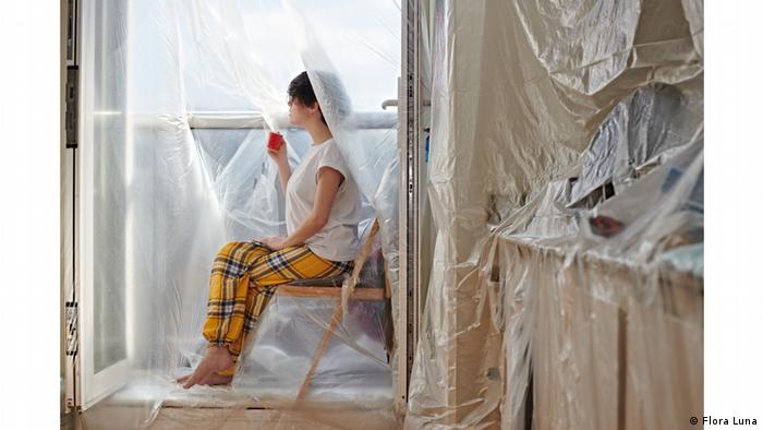 Photo from series Covered to D I S C O N N E C T: Girl sits on a wooden chair gazing through a plastic-covered window, the entire room covered in plastic as well (Flora Luna)