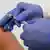 Pessoa com luvas azuis segura agulha de vacinação perto do braço de outra pessoa. Não é possível ver os rostos. 